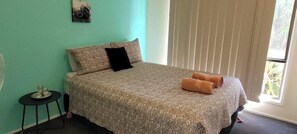 A bedroom with a bed, pillows, a small table with a lamp, and a window with blinds. - Roo Or 2 on Streeter (Agnes Water)