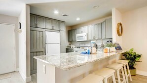 A kitchen with grey cabinets, a white refrigerator, a microwave, a sink, and a marble countertop. There are four white stools and a potted plant on the counter. - Community Perks! Sunny Hilton Head Island Retreat (Hilton Head Island)
