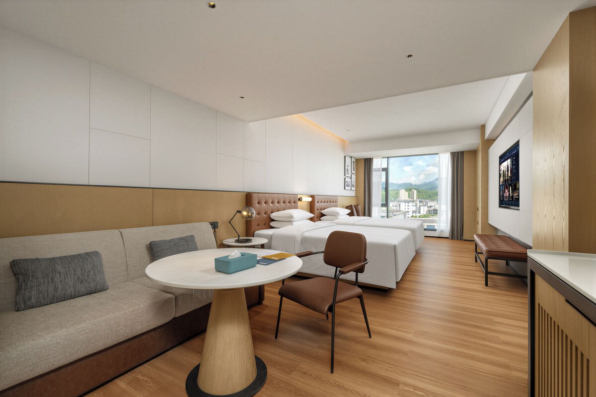 A modern hotel room with a sofa, a round table, a bed, and a view of the city.