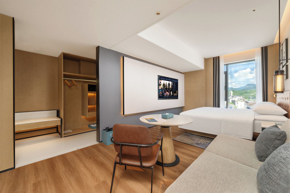 A modern hotel room with a bed, a seating area with a table and chair, a wardrobe, and a large window offering a city view.