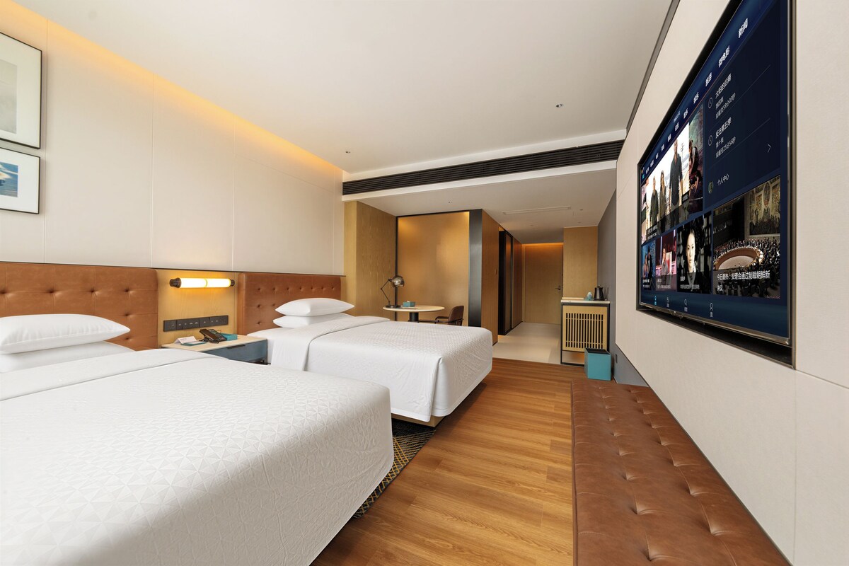 A hotel room with two beds, a desk, a chair, a TV, and a wall-mounted screen displaying content.
