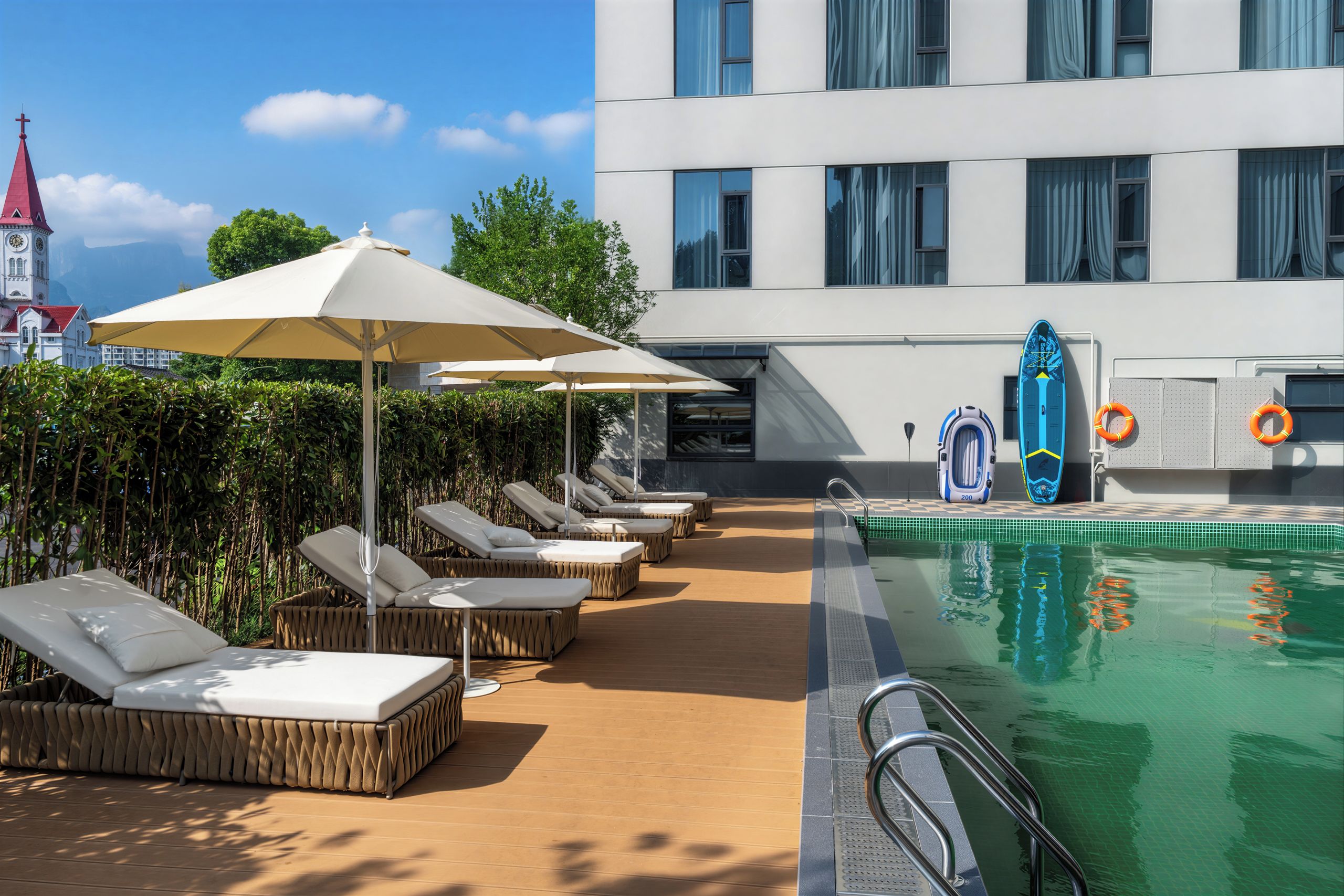 Zona junto a la piscina con sillones de descanso y sombrillas, piscina con boya salvavidas y un edificio con torre de reloj al fondo.