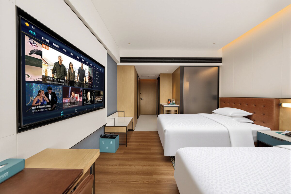 A hotel room with a large TV screen displaying movie posters, two beds, a bedside table, a nightstand, and a wooden floor.
