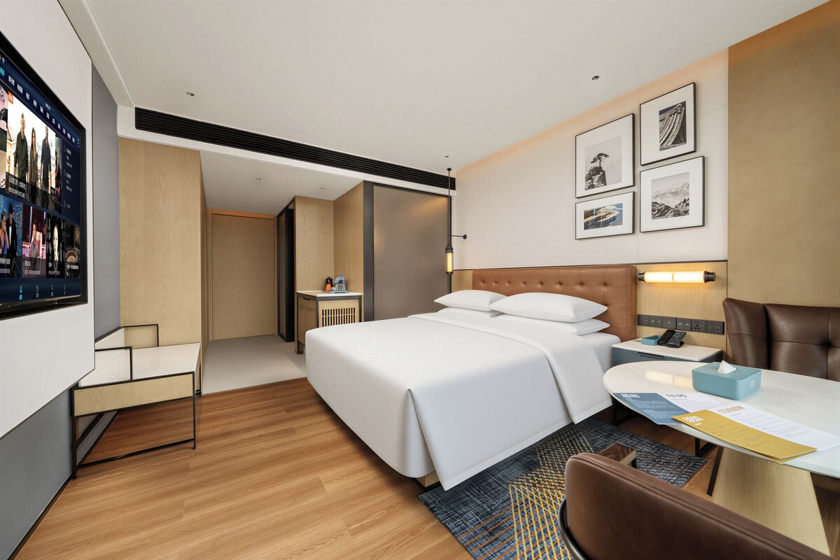 A modern hotel room with a large bed, a desk, a chair, and a television mounted on the wall.