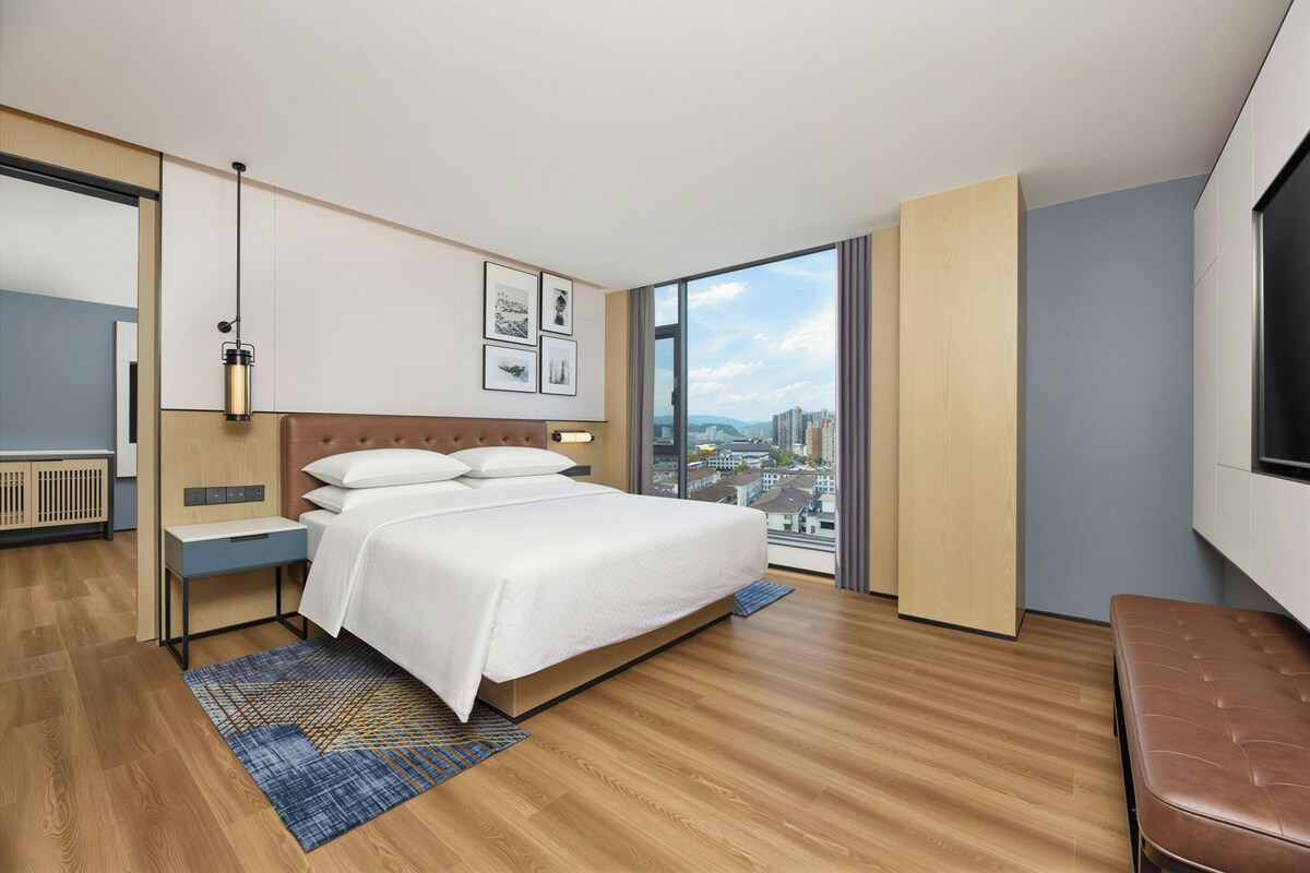 A modern hotel room with a large bed, a desk, a chair, a TV, and a city view.