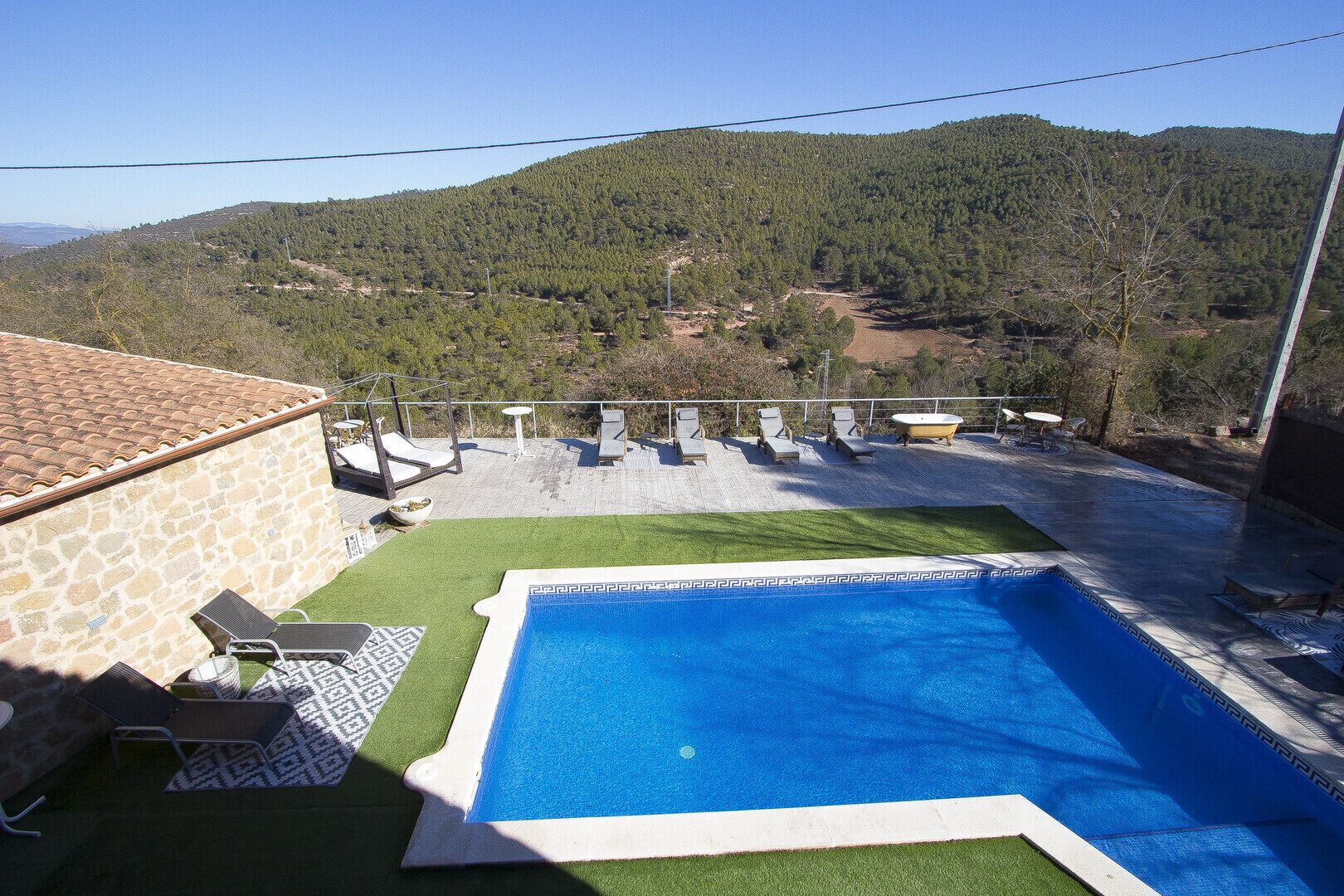 A swimming pool with a stone wall and lounge chairs, surrounded by a grassy area and a view of a forested hill.