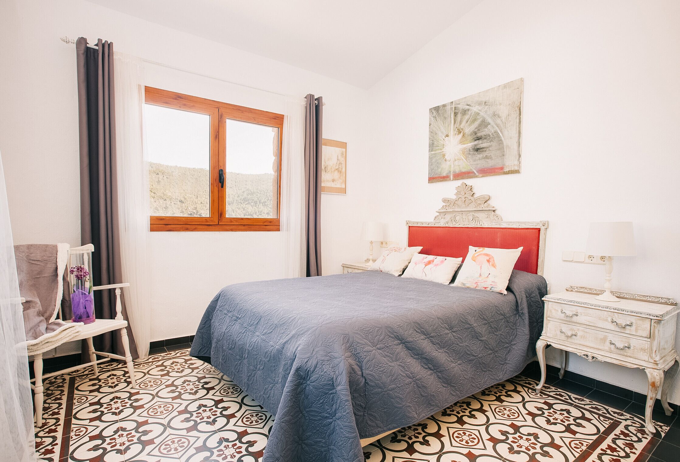 Un dormitorio con cama, mesita de noche, silla, ventana y un cuadro en la pared.
