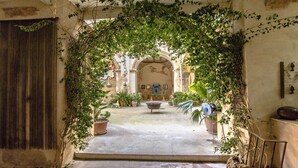 An arched doorway framed by lush greenery, leading to a courtyard with a fountain, potted plants, and a building with a mural. - Breathtaking 18th Century Converted Convent! (Vila-Rodona)