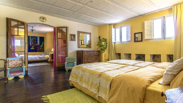 A room with a large bed, a dresser, a chair, and a mirror. There is a window with curtains and a door leading to another room.