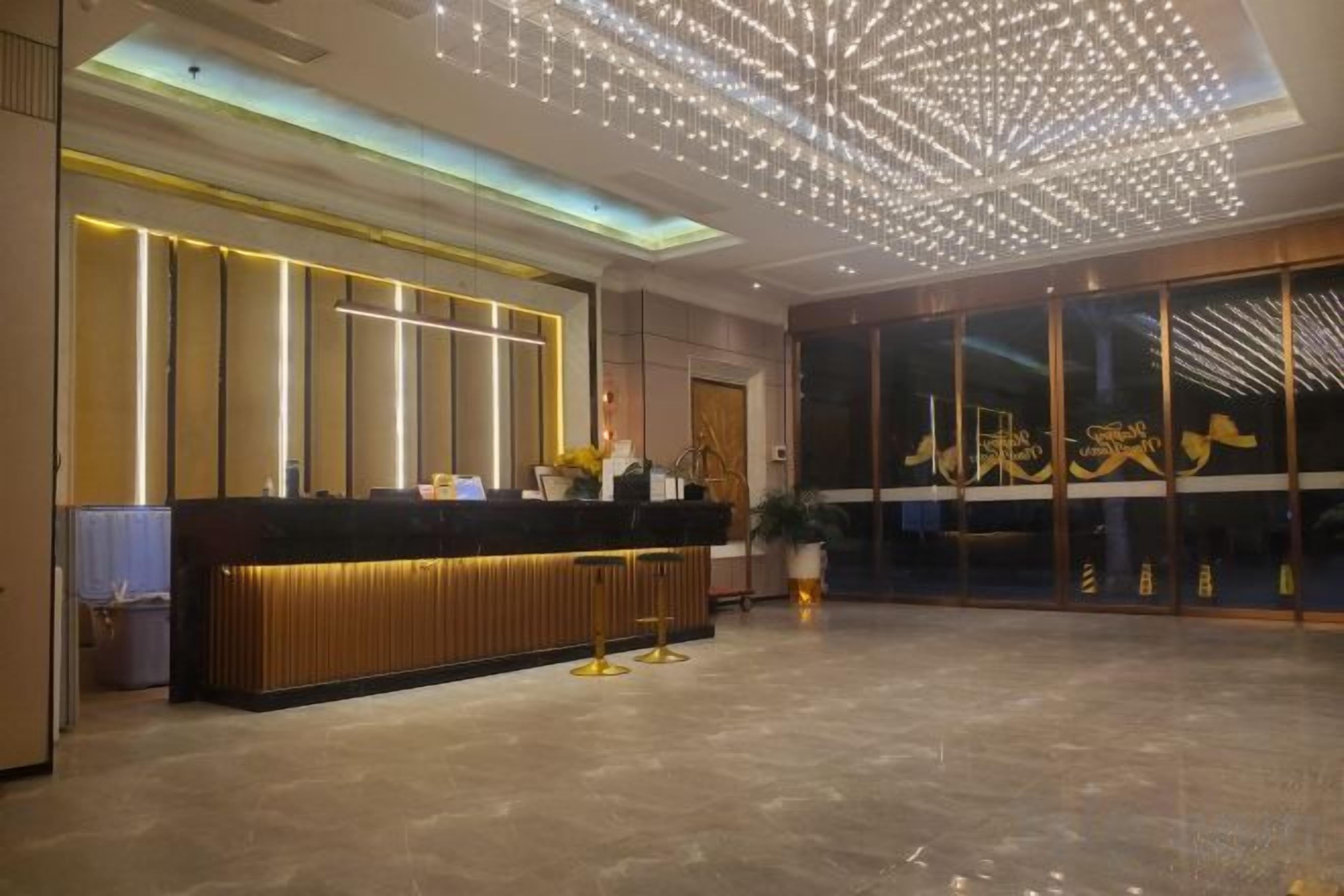 A hotel reception area with a modern design, featuring a reception desk, a large glass door with signage, and a ceiling with a geometric light fixture.