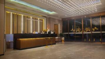 A hotel reception area with a modern design, featuring a reception desk, a large glass door with signage, and a ceiling with a geometric light fixture.