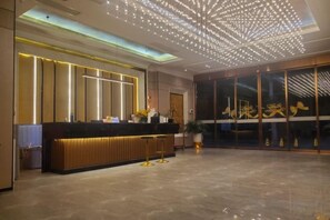 Zona de recepción de hotel con diseño moderno, que incluye un mostrador, una puerta de cristal grande con señalización y un techo con lámpara de diseño geométrico.