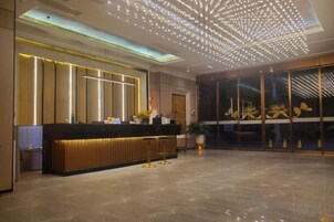 A hotel reception area with a modern design, featuring a reception desk, a large glass door with signage, and a ceiling with a geometric light fixture.