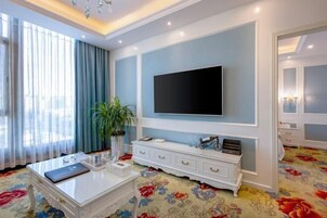 Habitación de hotel con una televisión de pantalla plana grande montada en la pared, una mesita consola blanca, una alfombra floral y un amplio ventanal con cortinas.