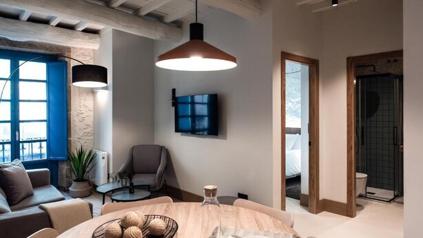 Un salon moderne avec une table ronde en bois, une fenêtre à cadre bleu, un fauteuil gris et un téléviseur fixé au mur.