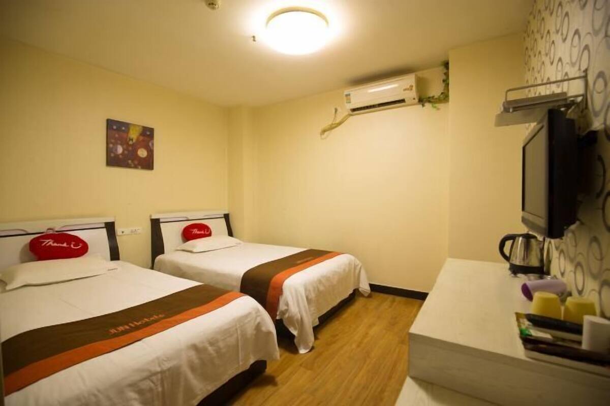 A hotel room with two beds, a desk, a TV, and an air conditioning unit.