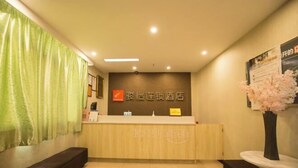 A hotel reception area with a wooden reception desk, a green curtain, a wall-mounted sign, and a vase with pink flowers. - Shushi Hotel (Shiyan)