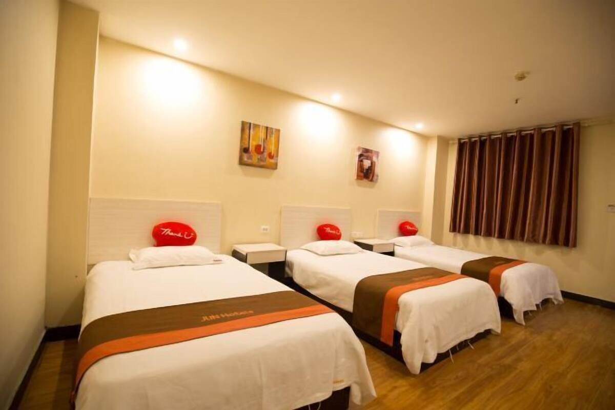 A hotel room with three beds, each with a red pillow and a white headboard. There are two wall-mounted artworks and a window with curtains.