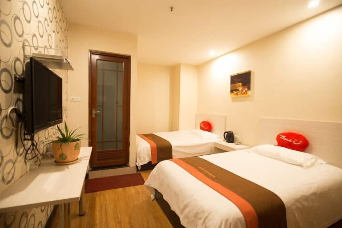 A hotel room with two beds, a TV, a plant, a door, and a small painting on the wall.