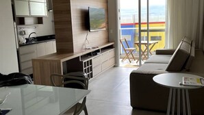 Una sala de estar moderna con un sofá, una mesa de centro y un televisor montado en la pared. Se observa una zona de cocina con alacenas y un lavabo.
