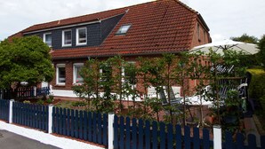 Et tosidet hus med rødt tegltag, hvid stakittence og en have med plantekasser samt en havestol på terrassen.