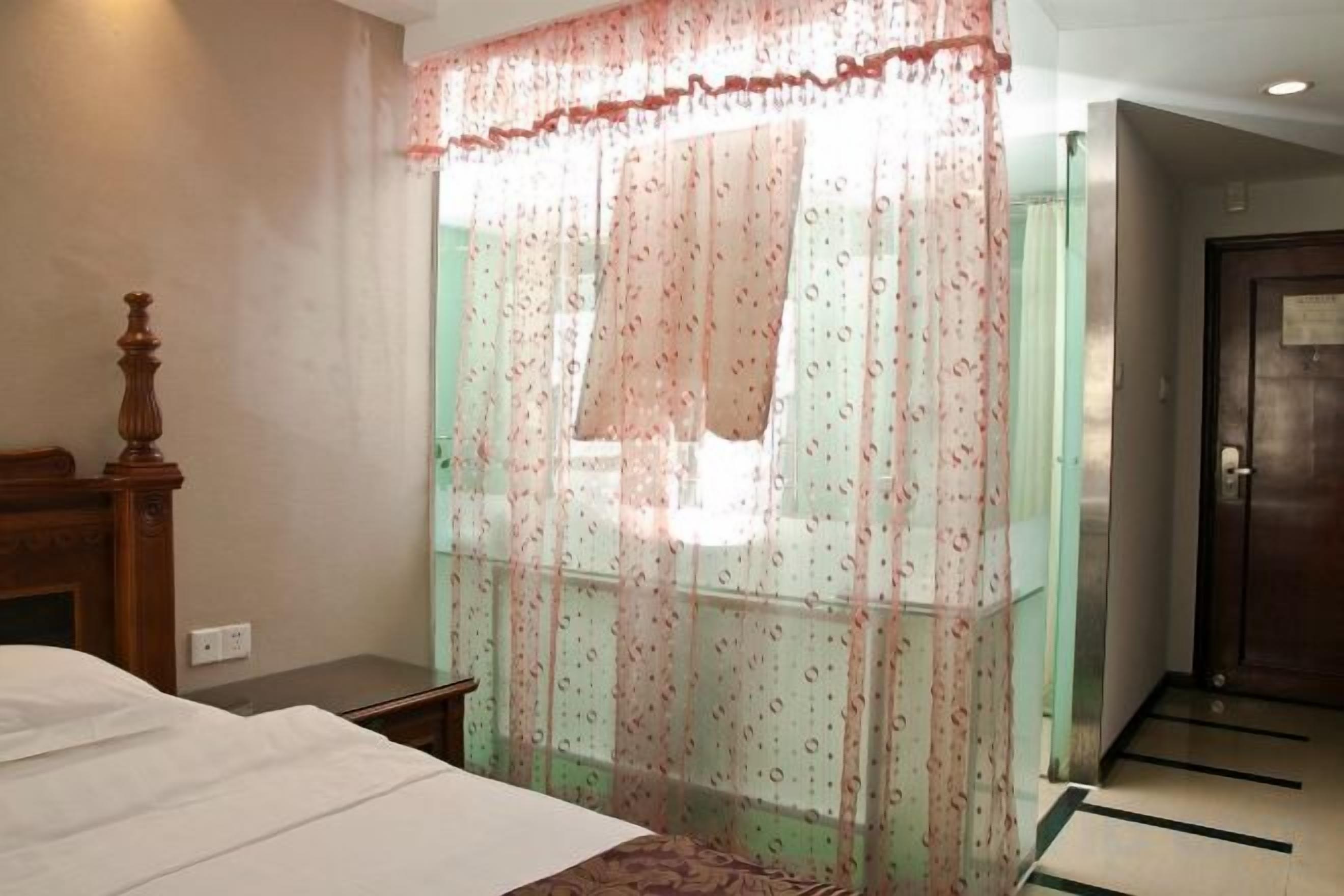 A hotel room with a bed, a wooden nightstand, a window with a sheer curtain, and a door.