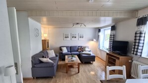 Un salón acogedor con un sofá gris, una mesa de centro de madera y un televisor sobre un mueble de madera. Hay tres cuadros enmarcados en la pared, una lámpara y una ventana con cortinas.