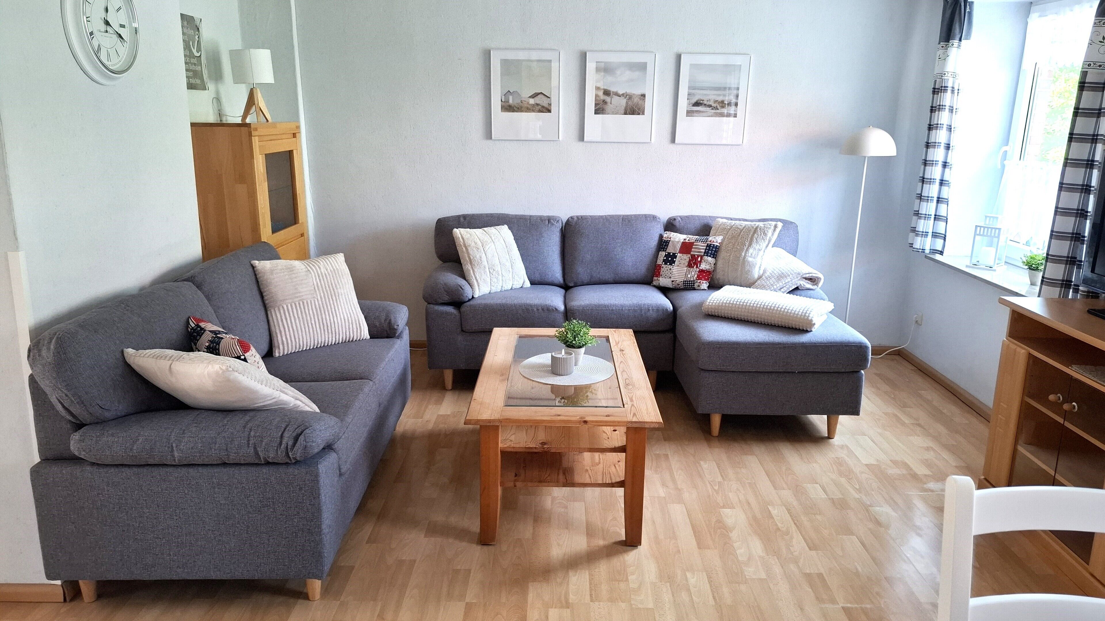 A modern living room with two grey sofas, a wooden coffee table, and a white chair. There are framed pictures on the wall and a lamp on a side table.