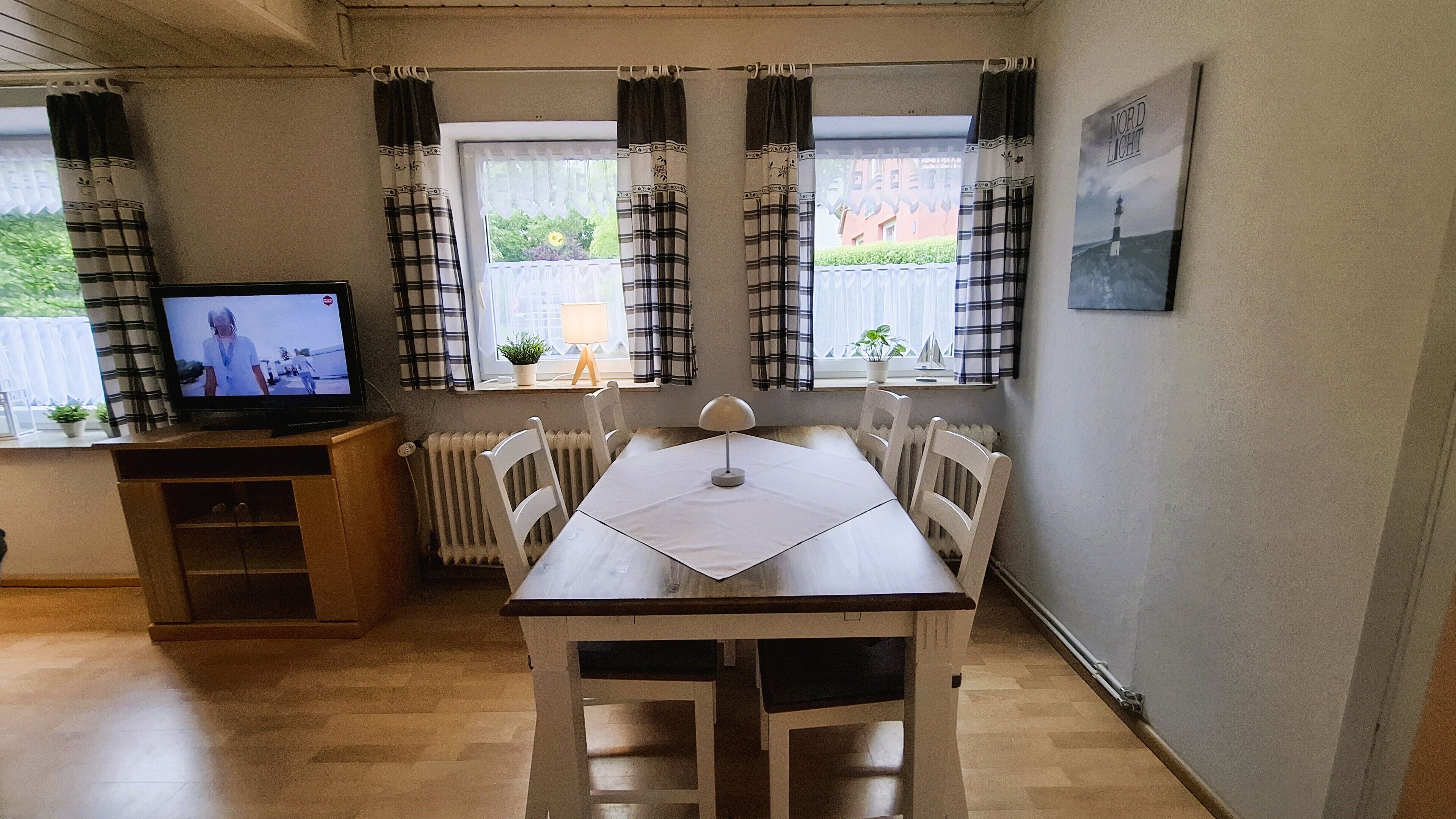 Een kamer met een houten tafel en stoelen, een televisie op een houten kast, en een raam met gordijnen.