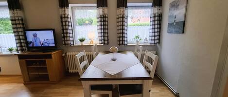 Een kamer met een houten tafel en stoelen, een televisie op een houten kast, en een raam met gordijnen.