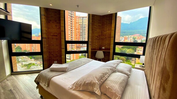 Ein modernes Schlafzimmer mit einem großen Bett, einem flachbildfernseher an der Wand und Blick auf die Stadt durch große Fenster.