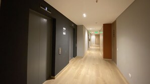 Ein Hotelgang mit Holzboden, einer dunklen Wand auf der einen Seite und einer hellen Wand auf der anderen. Es gibt Türen mit Hinweisschildern und einen Notausgang.
