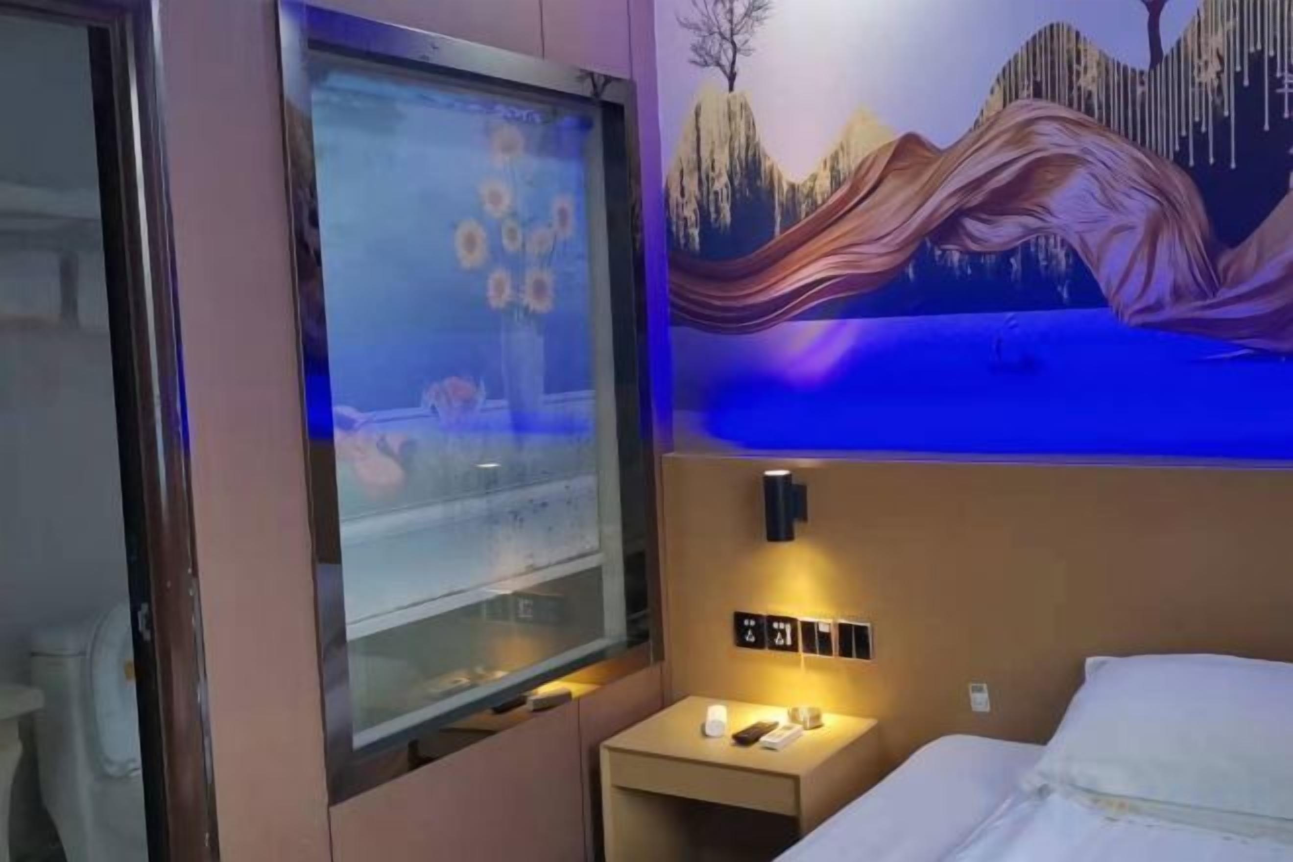 Una habitación de hotel con una cama, una mesita de noche con lámpara, un reloj y un televisor montado en la pared.
