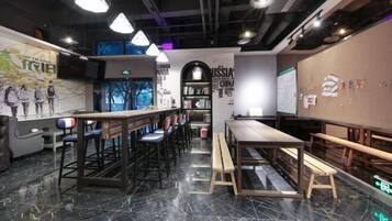Zona de bar moderna con mesas y taburetes altos, suelo de mármol y una pared con varios carteles y una estantería.