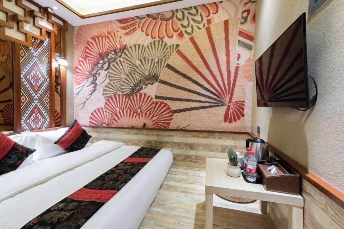 Ein Hotelzimmer mit einem Bett, einem wandmontierten Fernseher, einem kleinen Tisch mit einer Vase und einer Flasche sowie einem dekorativen Wandteppich.