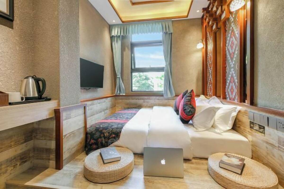 Ein Hotelzimmer mit einem Bett, zwei Nachttischen mit Büchern, einem Laptop, einem Fernseher und einem Fenster mit Vorhängen.