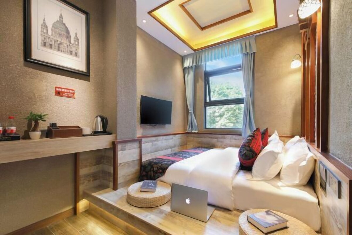 A hotel room with a bed, a laptop, a TV, a window with curtains, a framed picture on the wall, and a small table with a plant and a book.