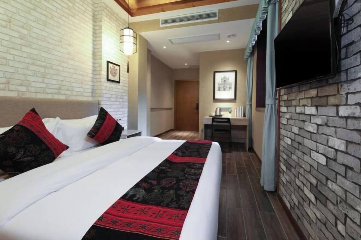 Ein Hotelzimmer mit Backsteinwand, einem Bett mit roten und schwarzen Kissen und einer Bettdecke mit Muster, einem an der Wand montierten Flachbildfernseher und einer Holzbalkendecke.