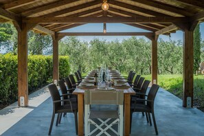A covered outdoor dining area with a long table set for a meal, surrounded by chairs.