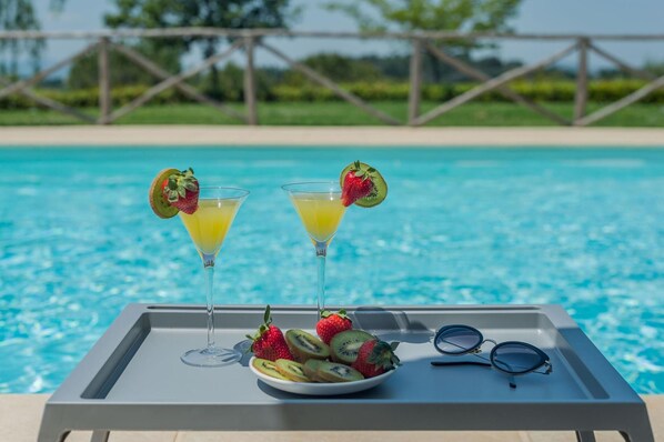 A poolside tray with two drinks, a bowl of fruit, and a pair of sunglasses.
