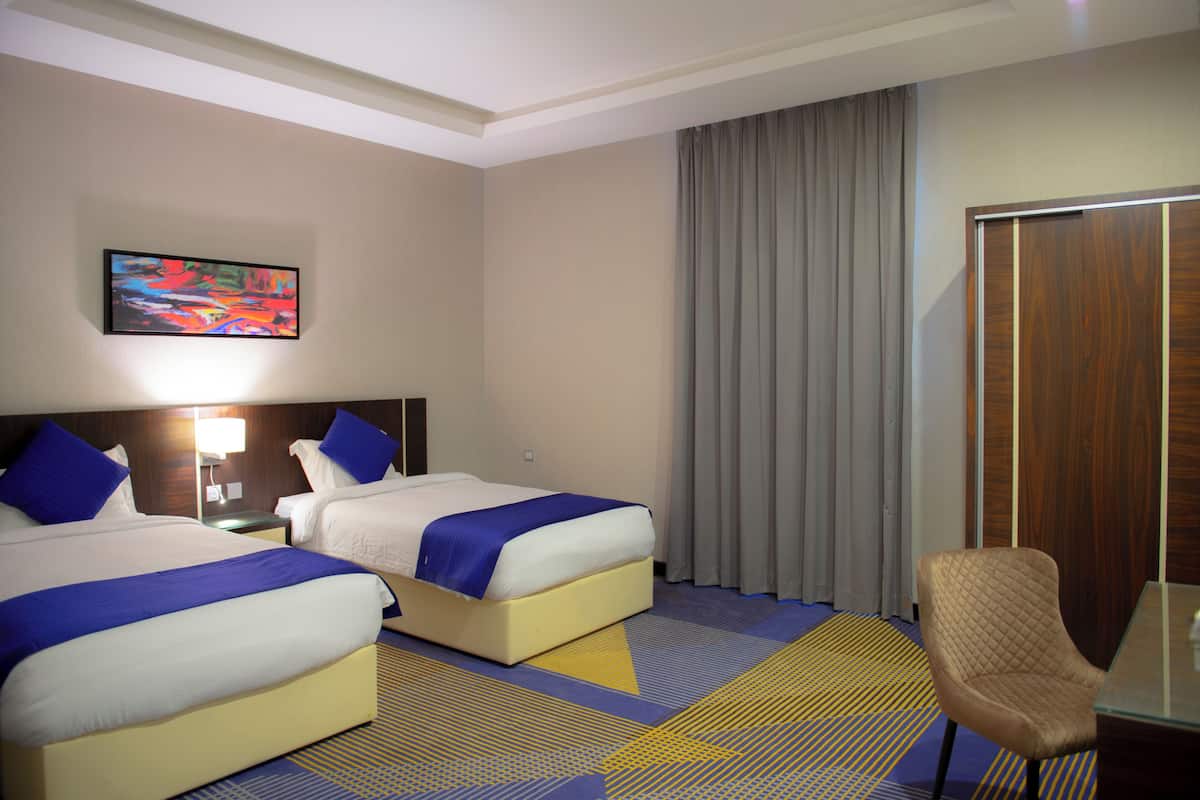 A hotel room with two beds, a desk, a chair, a painting on the wall, and a window with curtains.