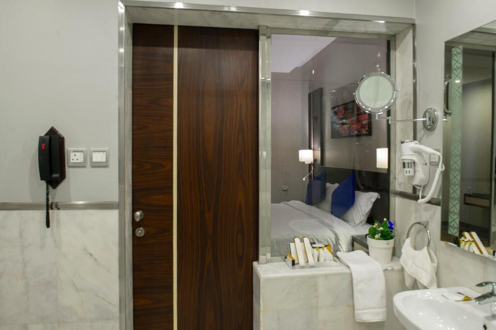 A modern bathroom with a marble floor, a wooden door, a bed with a blue pillow, a white sink, and a mirror.