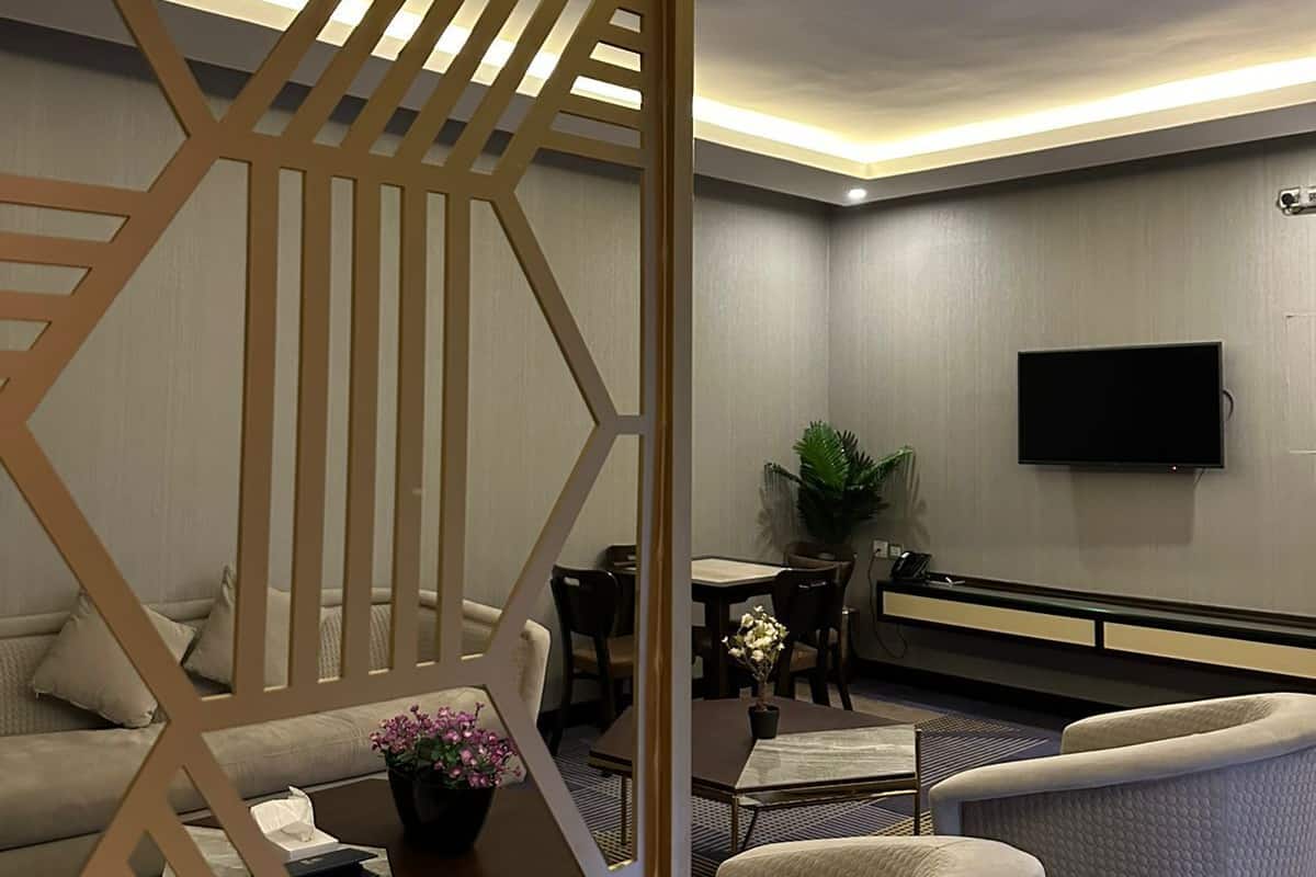 A modern hotel room with a seating area, a flat-screen TV mounted on the wall, and a decorative partition.