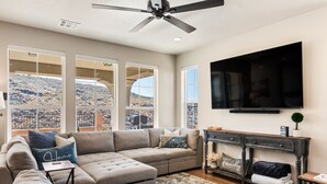 A spacious living room with a large sectional sofa, a flat-screen TV mounted on the wall, and a view of a mountainous landscape through large windows. - Splash Pad: Pickleball, E-bike, Scooters, Bicycles (Washington)