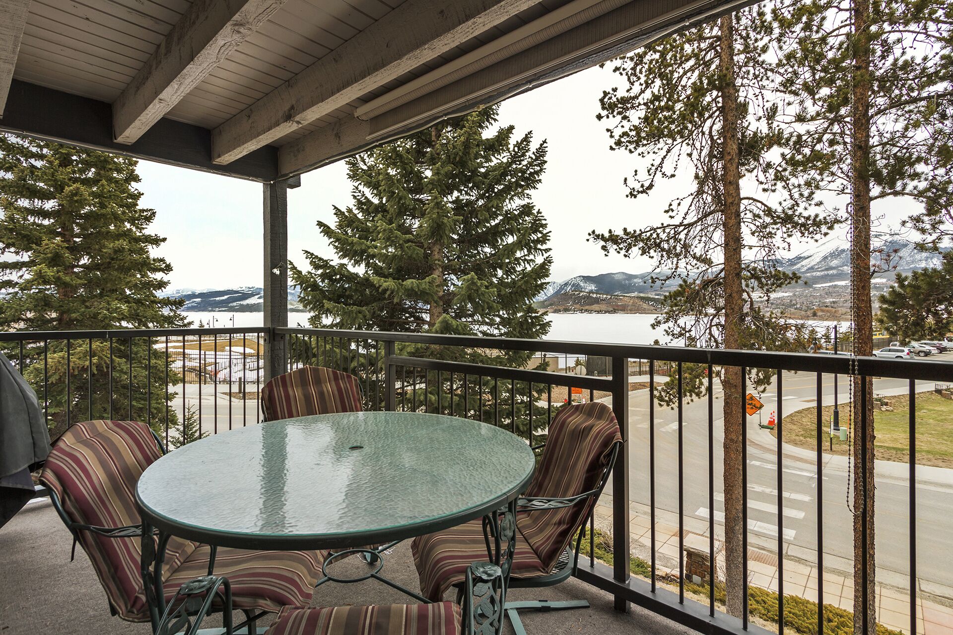 Een balkon met een glazen tafel en gestreepte stoelen, met uitzicht op een weg en een meer met bergen in de verte.