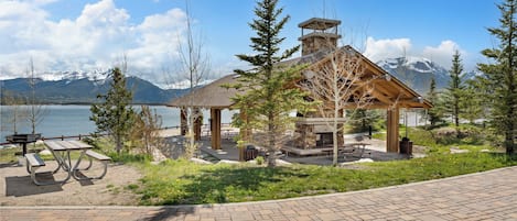 A lodge with a stone chimney, picnic tables, and a view of a lake and mountains.