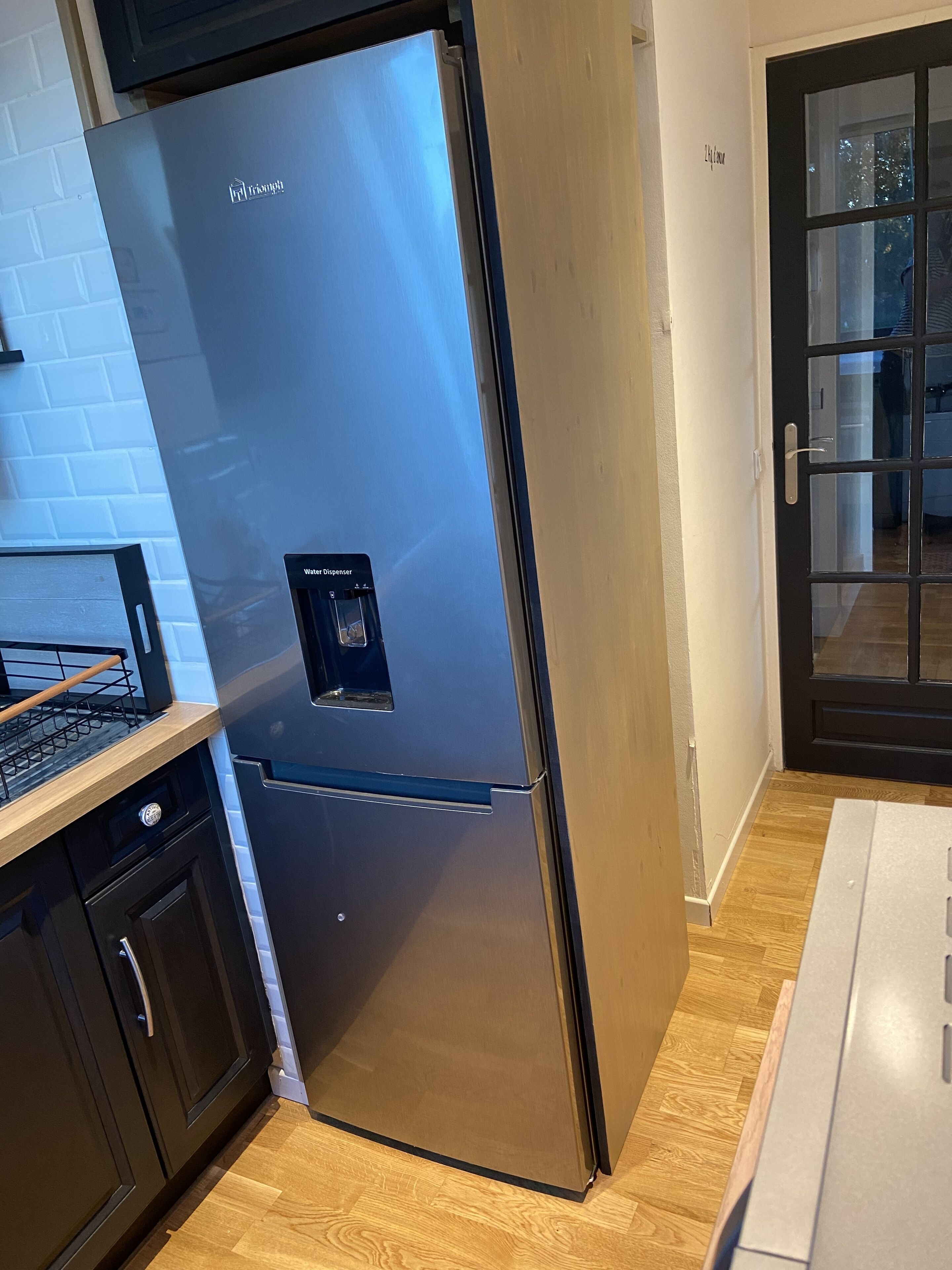 A stainless steel refrigerator with a water dispenser, located in a kitchen with wooden flooring and a white brick backsplash.