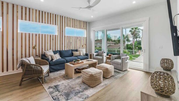 Ein modernes Wohnzimmer mit einer Couch, einem Couchtisch und Hocker. Es gibt einen Deckenventilator und ein großes Fenster mit Blick auf Palmen.
