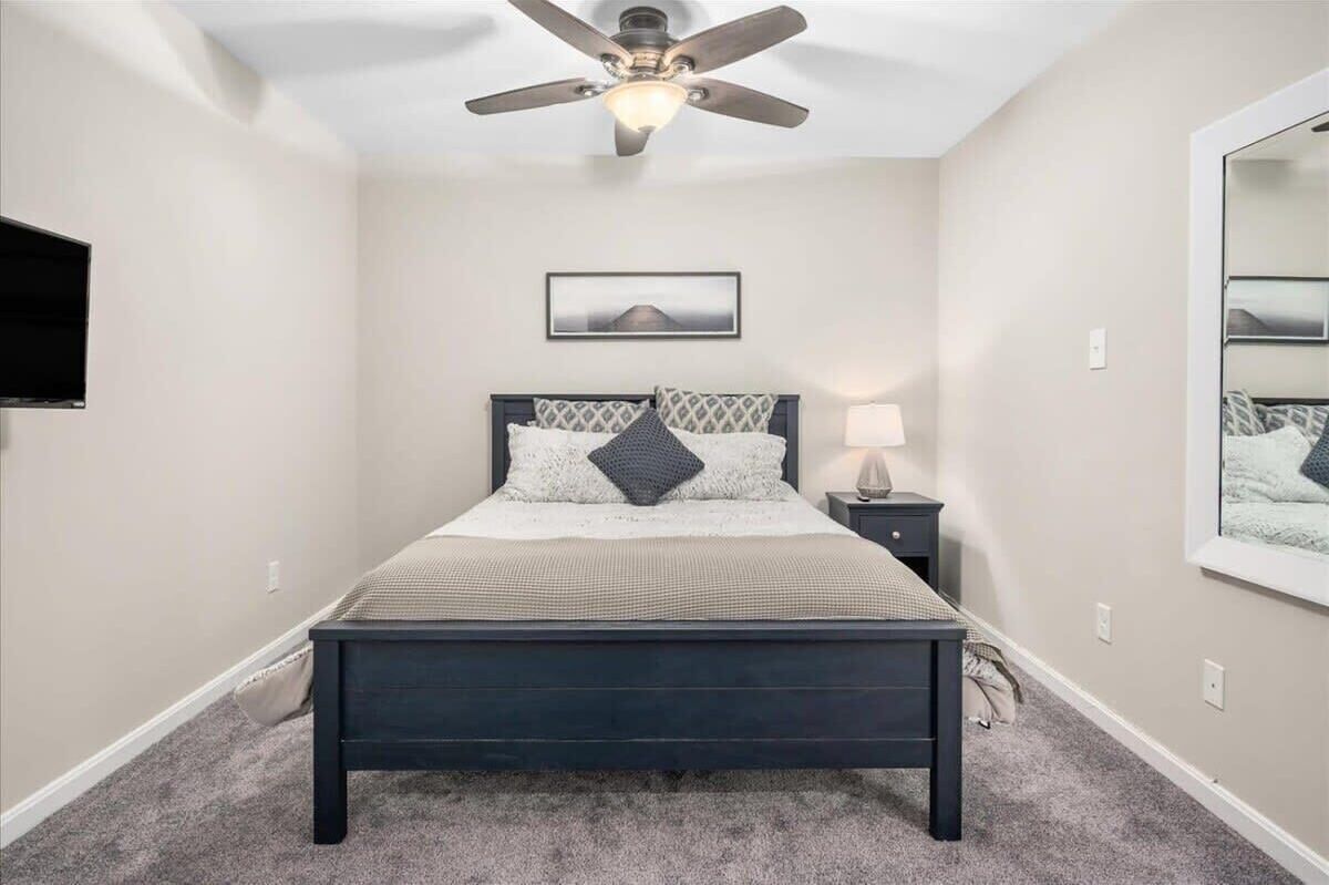 A bedroom with a bed, a ceiling fan, a nightstand, a lamp, a framed picture, and a mirror.
