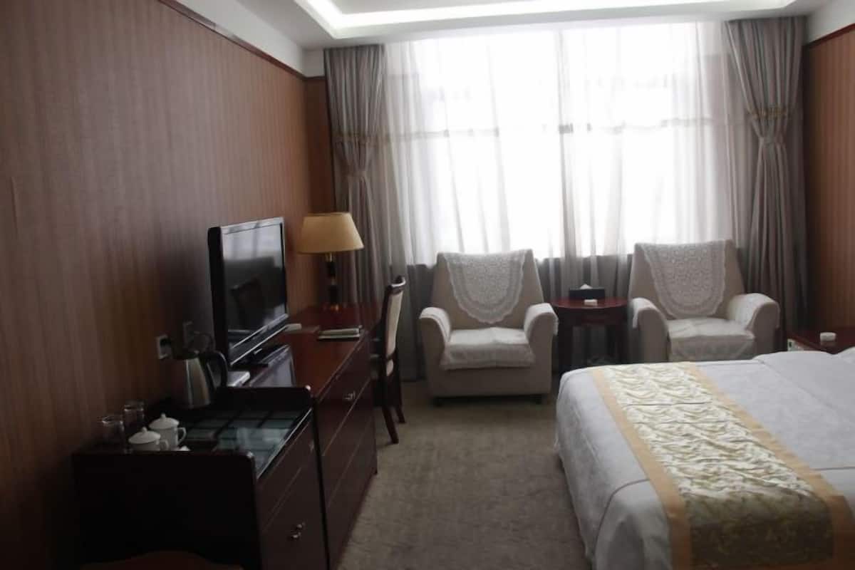 A hotel room with a bed, two armchairs, a TV, a desk, and a window with curtains.